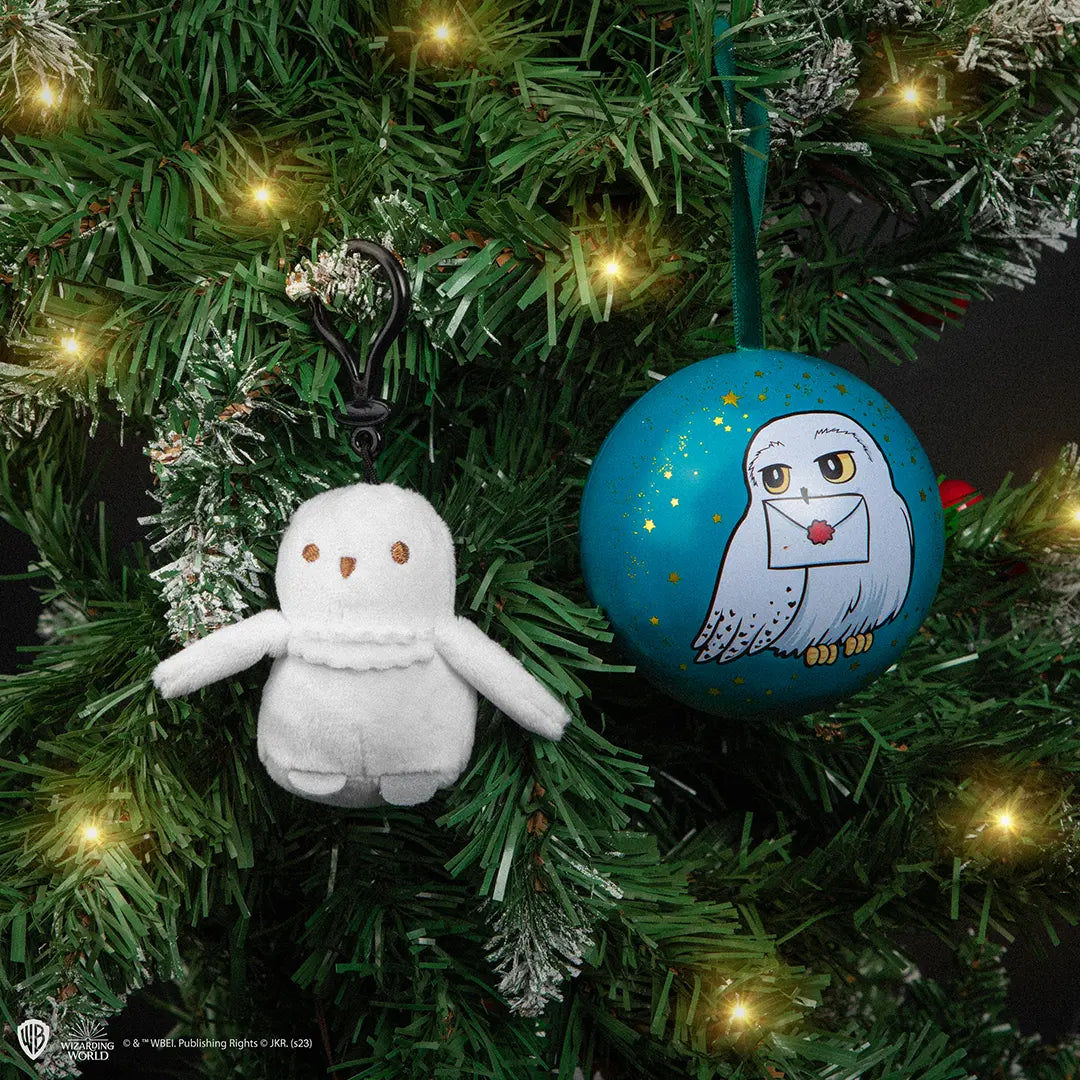 Witte pluchen Hedwig-sleutelhanger met goudbruine ogen en snavel, gespreide vleugels en een zwarte clip. Hij hangt tussen groene kersttakken met lichtjes, naast een turquoise kerstbal met gouden sterren en een uil met een brief.