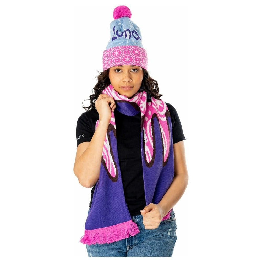 Model met een lichtblauwe gebreide muts met paarse tekst ‘Luna’, een roze-witte omslag en roze pompon. Ze draagt een bijpassende roze-paarse sjaal, houdt die met beide handen vast en staat tegen een witte achtergrond.