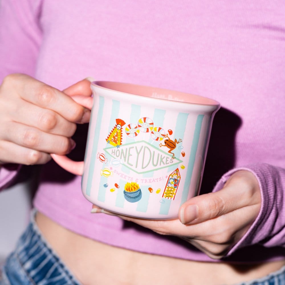 Roze keramieken mok met mintgroene strepen, Honeydukes-opschrift en kleurrijke snoepillustraties. Een persoon houdt de mok met beide handen vast, één aan het oor en één onder de bodem, tegen een roze-paarse trui.