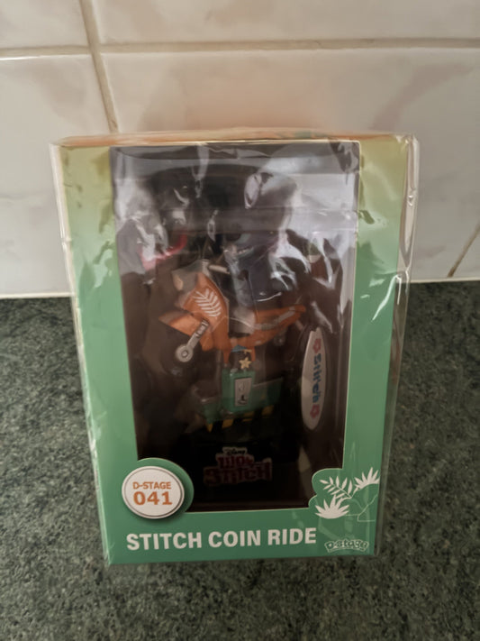 Blauwe Stitch-figuur op een oranje raket in een mintgroene kartonnen doos met doorzichtig plastic venster en de tekst ‘Stitch Coin Ride’. De doos staat op een groen gevlekt aanrecht tegen witte tegels.