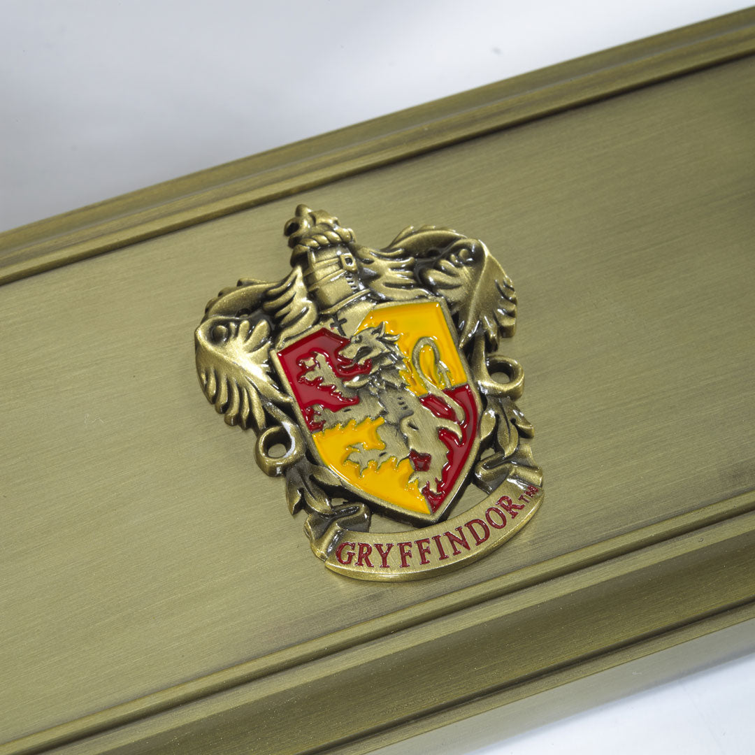 Schuin gefotografeerde, bronskleurige toverstokhouder met metaalachtige afwerking en opstaand Griffoendorwapen: een leeuw op een rood-geel schild, met daaronder ‘GRYFFINDOR’. De rechthoekige houder ligt op een witte achtergrond.