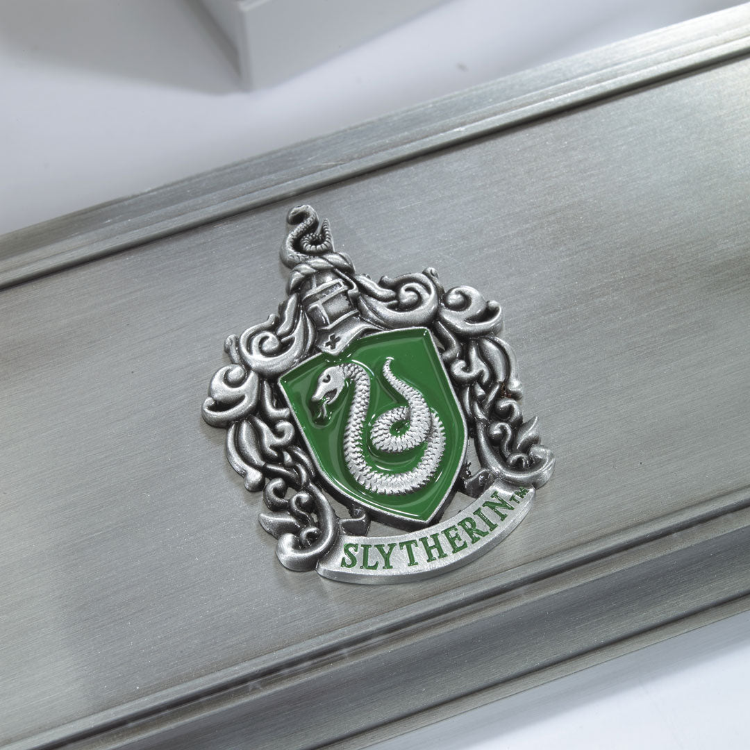 Detail van een schuin liggende, zilverkleurige toverstokhouder met geborstelde afwerking. Op de witte achtergrond toont de houder een groen wapenschild met zilverkleurige slang, sierkrullen en het opschrift ‘SLYTHERIN’.