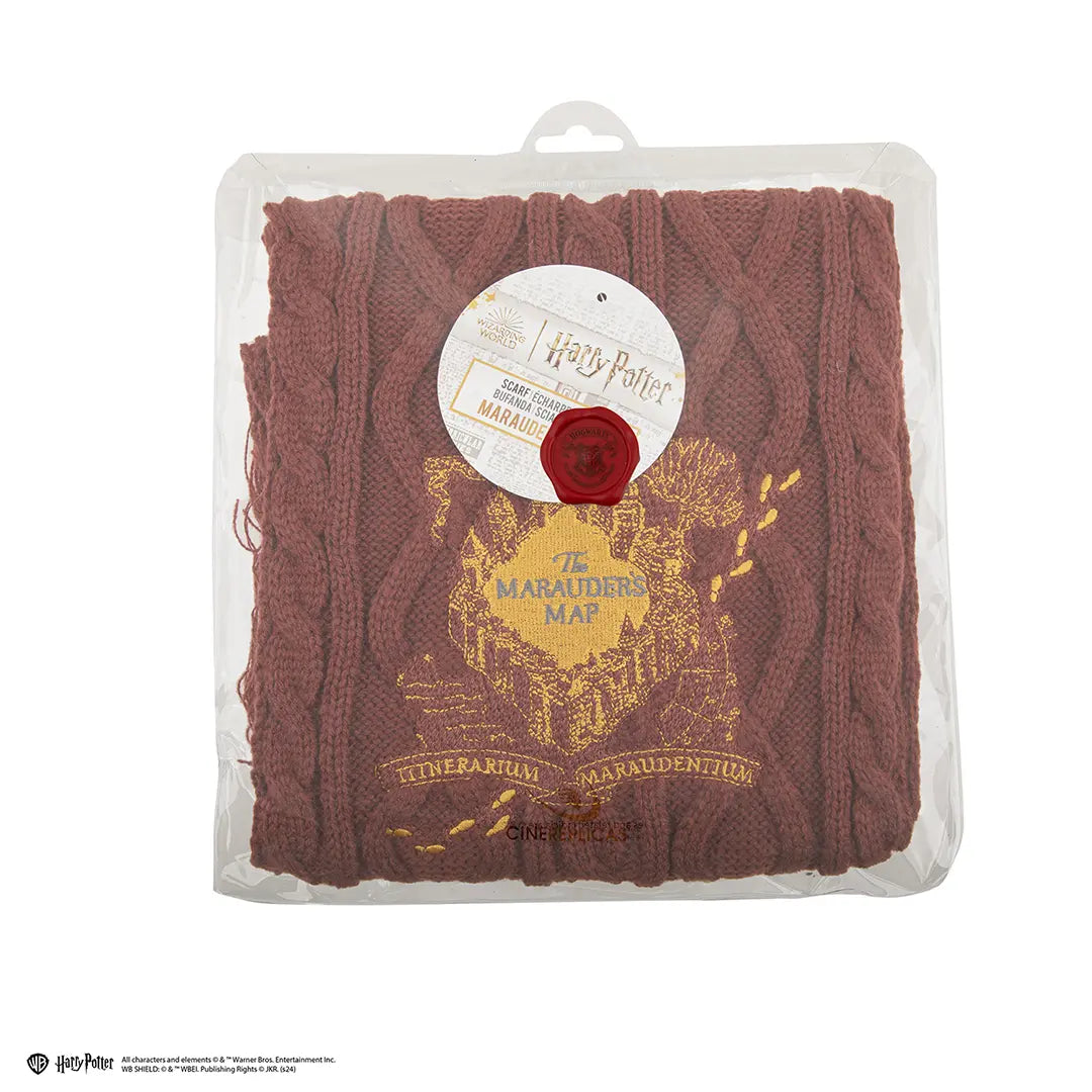 Opgevouwen bruinrode, gebreide sjaal met kabelpatroon en goudgele Sluipwegwijzer-opdruk met voetstappen. In een doorzichtige kunststof verpakking met rond Harry Potter-label, tegen een witte achtergrond.