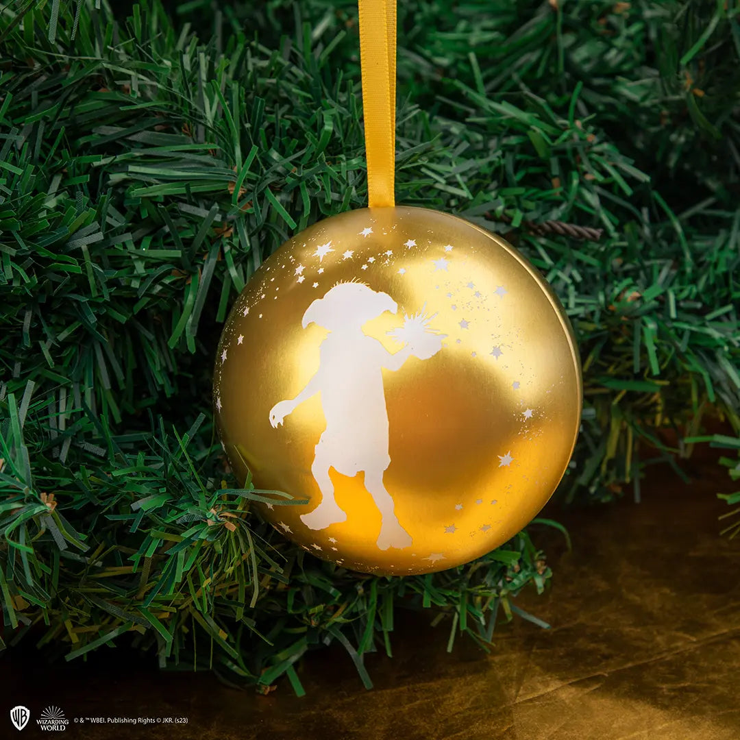 Ronde, goudkleurige metalen hanger met een wit silhouet van Dobby met geheven hand, omringd door witte sterretjes. De hanger hangt aan een geel lint tussen groene kerstboomtakken, boven een bruine ondergrond.