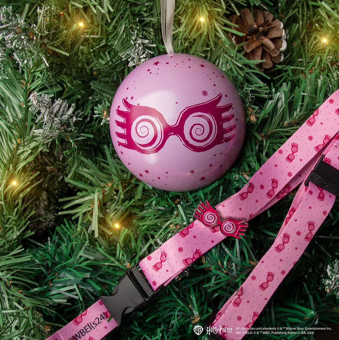 Roze stoffen keycord met brilletjes, sterren, zwarte kunststof sluiting en metalen brillenpin, schuin tussen groene kersttakken. Ernaast hangt een roze kerstbal met Luna’s bril, omringd door lichtjes en dennenappels.