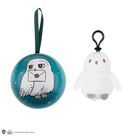 Witte pluchen Hedwig-sleutelhanger, rechtop met gespreide vleugels, grijze voetjes en zwarte clip. Links een turquoise bol met gouden sterren, een afbeelding van Hedwig met een brief en een groen ophanglint, op een witte achtergrond.