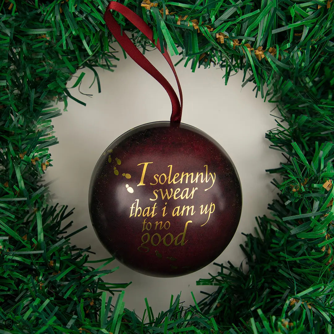 Ronde, glanzende bordeauxrode capsule met gouden voetstappen en de tekst ‘I solemnly swear that i am up to no good’. Hangt aan een bordeauxrood stoffen lint in een groene kunstkerstkrans tegen een gebroken witte achtergrond.