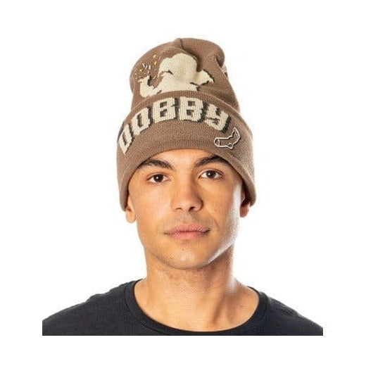 Een model met zwart shirt draagt een bruine gebreide muts met een beige afbeelding van Dobby, de tekst ‘DOBBY’ en een sokje op de omslag. Het model kijkt recht vooruit tegen een witte achtergrond.