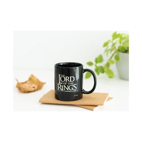 Zwarte, glanzende keramische mok met wit The Lord of the Rings-logo en het oor rechts, op bruin papier. Op de witte achtergrond liggen links droge bladeren en staat rechts een groene plant in een lichte pot.
