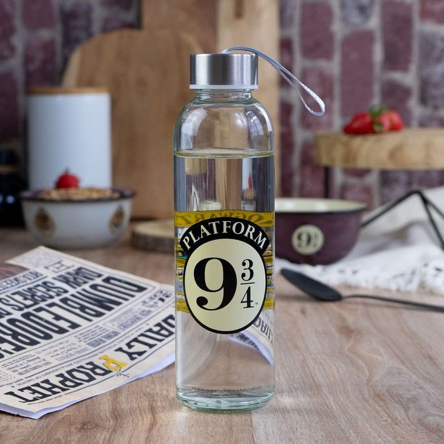 Transparante glazen drinkfles met zilverkleurige dop, grijze draaglus en zwart-geel ‘Platform 9¾’-embleem, rechtop op een houten tafel. Op de onscherpe achtergrond staan kommen en snijplanken tegen een bakstenen wand.