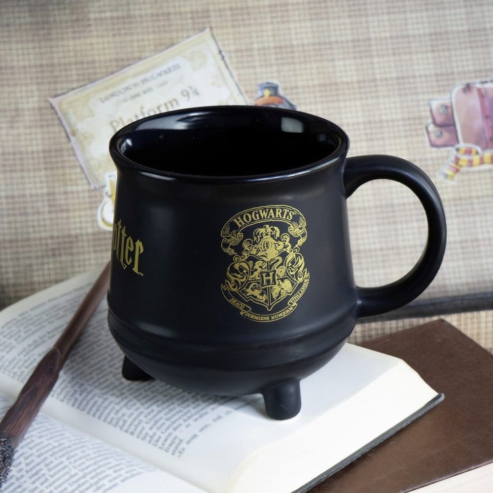 Zwarte keramische ketelmok met een goudkleurig Zweinsteinwapen, korte pootjes en een oor rechts, op een opengeslagen boek. Ernaast ligt een bruine toverstok; de beige geruite achtergrond bevat Harry Potter-illustraties.