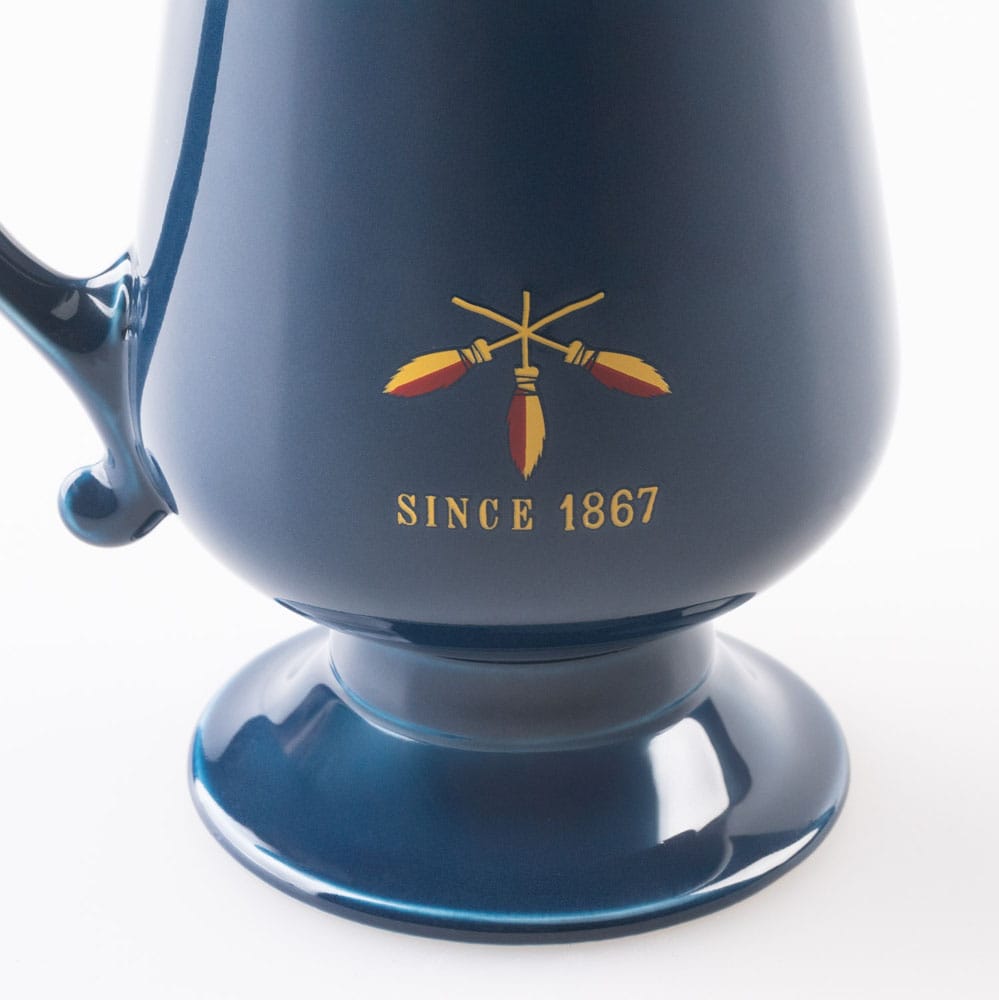 Detail van een glanzende donkerblauwe keramieken boterbierbeker op ronde voet, met links een sierlijk oor. Op de beker staan drie gekruiste bezems in geel en rood en de tekst ‘SINCE 1867’, tegen een witte achtergrond.