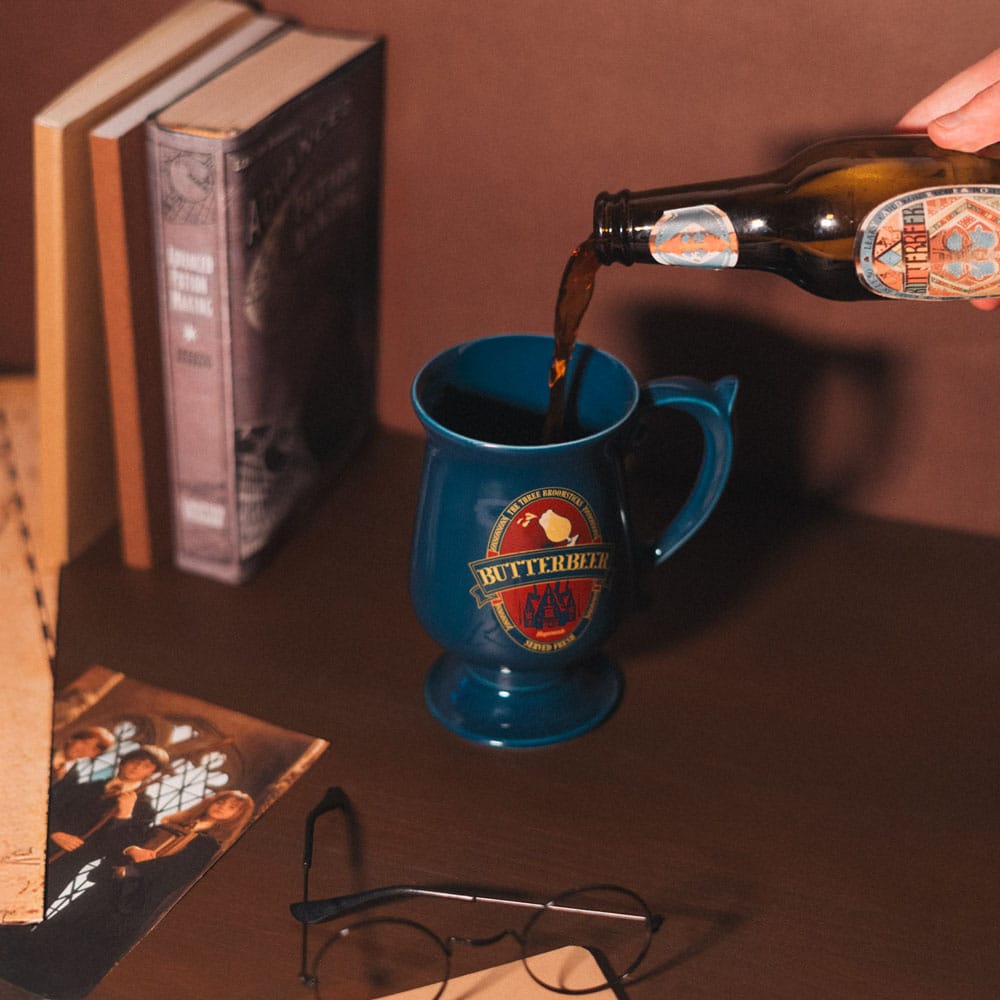 Glanzende donkerblauwe keramische beker op voet met oor en rood-goudkleurig Butterbeer-logo. Een hand schenkt uit een bruine fles in de beker, die op een bruine tafel staat met boeken, een foto en een ronde bril.