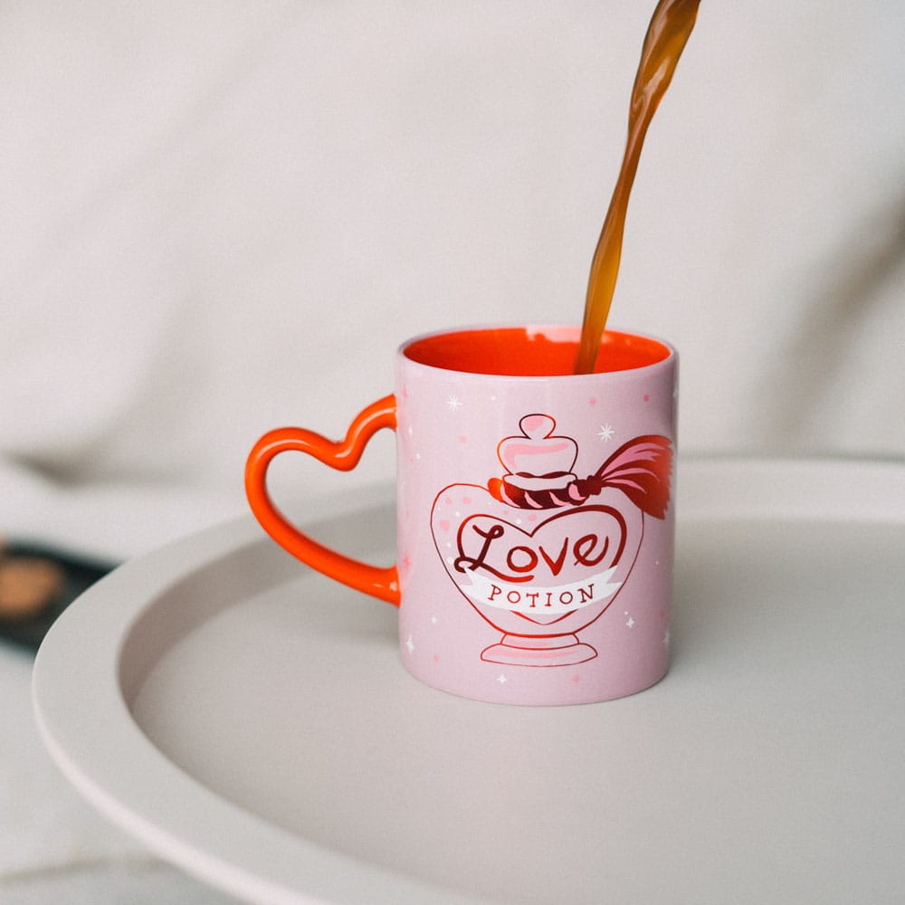 Roze keramieken mok met rood hartvormig oor, rode binnenkant en een flesje met de tekst ‘Love Potion’ op de voorkant. Bruine vloeistof wordt in de mok geschonken, die op een licht dienblad staat tegen een lichte achtergrond.