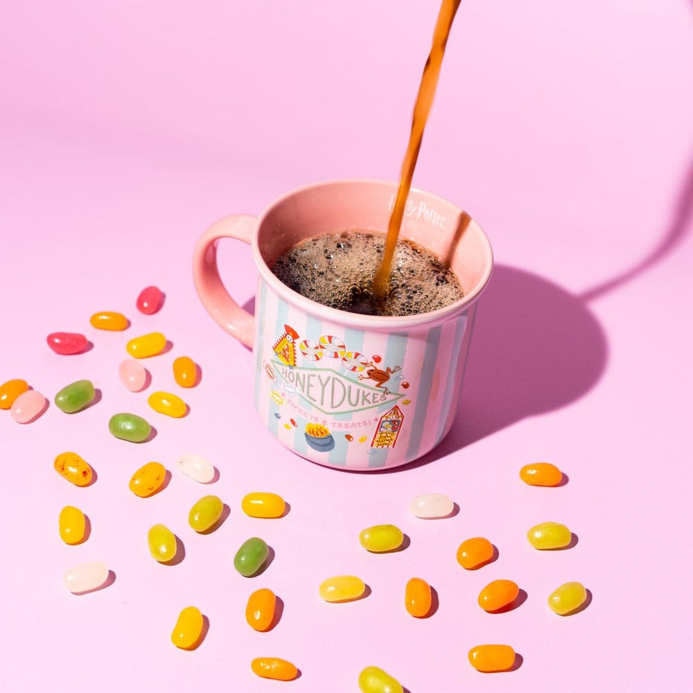 Keramieken Honeydukes-mok met roze en lichtblauwe strepen, snoepillustraties en het oor links. Er wordt een bruine drank ingeschonken; de mok staat tussen kleurrijke snoepboontjes op een roze ondergrond met roze achtergrond.