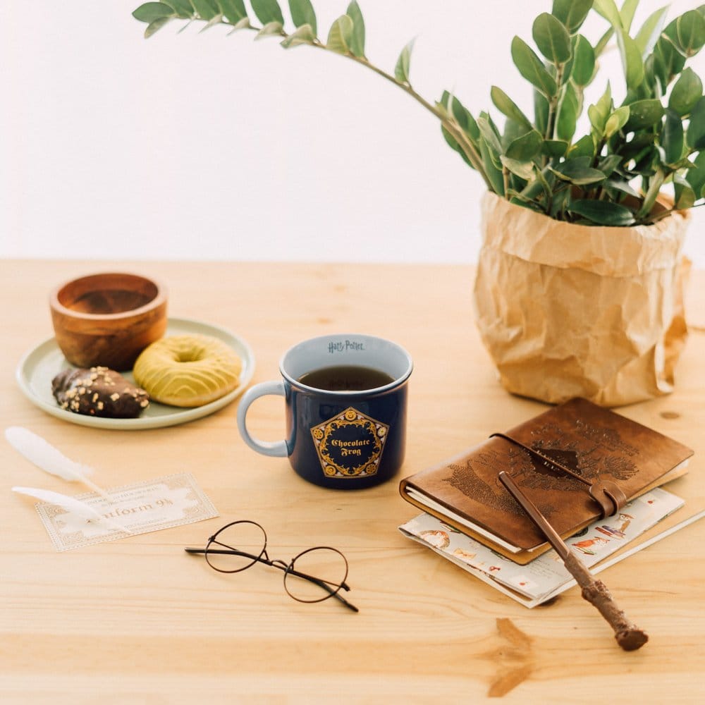Donkerblauwe keramische mok met lichtblauw oor links en goudkleurig Chocolate Frog-embleem, gevuld met donkere drank. Op een houten tafel met gebak, ronde bril, notitieboek, toverstok en groene plant, tegen een lichte muur.