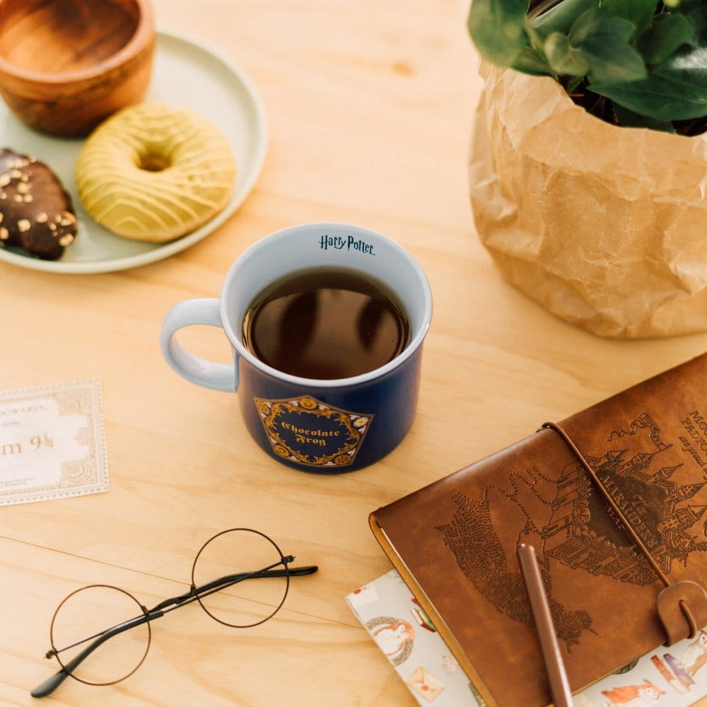 Donkerblauwe keramische mok met goudkleurig Chocokikker-embleem, lichtblauwe binnenkant en oor links, gevuld met een donkere drank. Op een licht houten tafel met gebak, een plant, een ronde bril en een bruin notitieboek.
