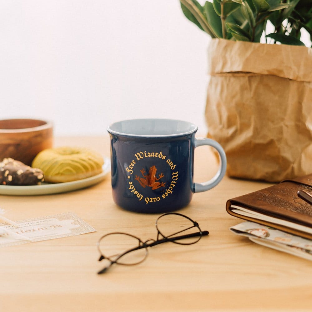 Donkerblauwe keramieken mok met lichtblauw oor rechts, een bruine kikker en goudkleurige tekst. De mok staat op een houten tafel met een bril, boeken en gebak, met een plant in een bruine papieren zak op de achtergrond.