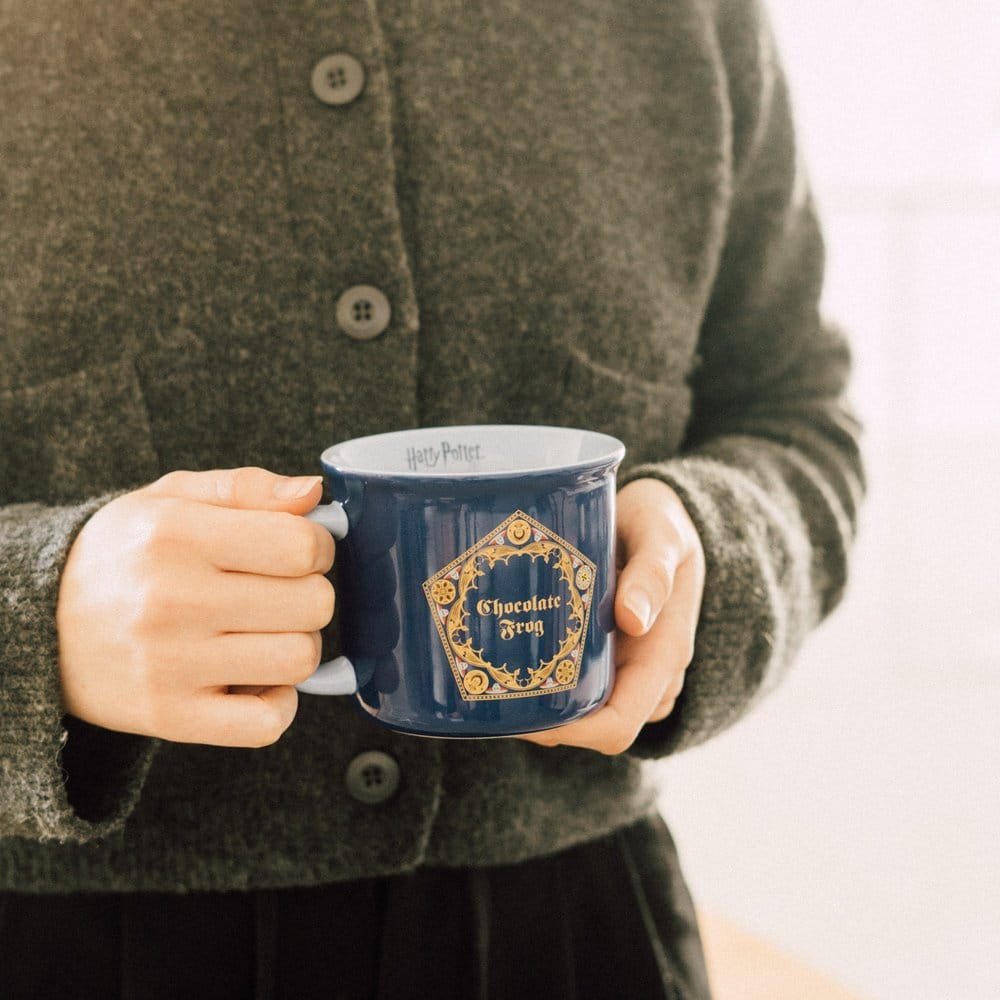 Glanzende donkerblauwe keramische mok met witte binnenkant en goudkleurig ‘Chocolate Frog’-embleem. Een persoon in een grijsgroen gebreid vest houdt de mok met twee handen vast, met het oor links, tegen een lichte achtergrond.