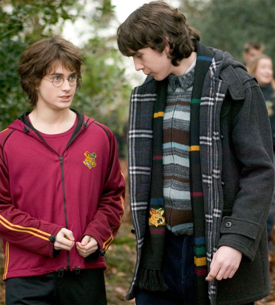 Zwarte gebreide sjaal met rode, gele, groene en blauwe strepen, Hogwarts-wapen en franjes, los om de hals van een man in een donkere jas. Hij staat naast Harry Potter, met bomen en mensen op de onscherpe achtergrond.