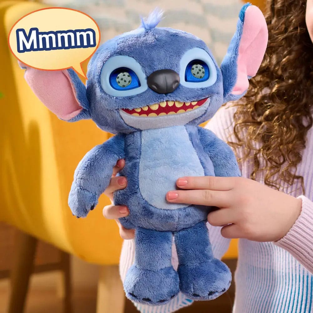 Blauwe pluchen Stitch met lichtblauwe buik, grote oren met roze binnenkant en een brede grijns. Een kind houdt de knuffel rechtop en drukt op zijn buik, met een gele stoel op de achtergrond en linksboven een tekstballon met ‘Mmmm’.