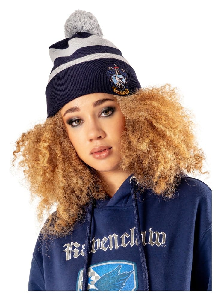 Donkerblauwe gebreide Ravenklauw-muts met lichtgrijze strepen, grijze pompon en geborduurd wapen op de omslag. Een model met blonde krullen en een blauwe hoodie draagt de muts met het hoofd licht schuin tegen een witte achtergrond.