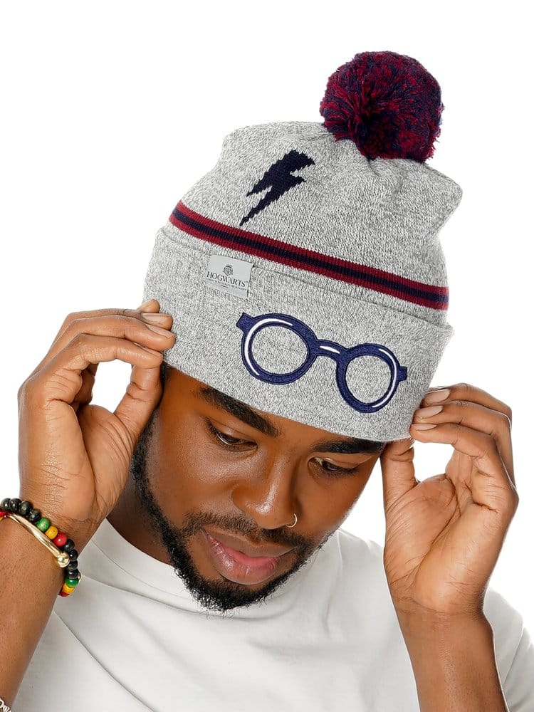 Grijze gebreide Harry Potter-muts met rood-donkerblauwe pompon en strepen, een donkerblauwe bliksemschicht en bril op de omslag. Een man kijkt omlaag en houdt de muts met beide handen vast tegen een witte achtergrond.