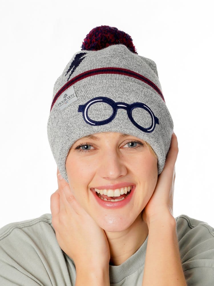 Grijze gebreide Harry Potter-muts met donkerblauwe bril, bliksemschicht, rood-blauwe strepen en pompon. Een glimlachende persoon draagt de muts met beide handen langs het gezicht, tegen een witte achtergrond.