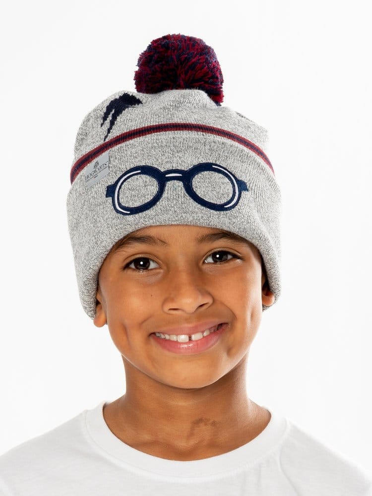 Een glimlachend kind draagt een grijze gebreide Harry Potter-muts met een donkerblauwe bril op de omslag, een bliksemschicht, rode en blauwe strepen en een rood-blauwe pompon. Het kind draagt een wit shirt en staat voor een witte achtergrond.