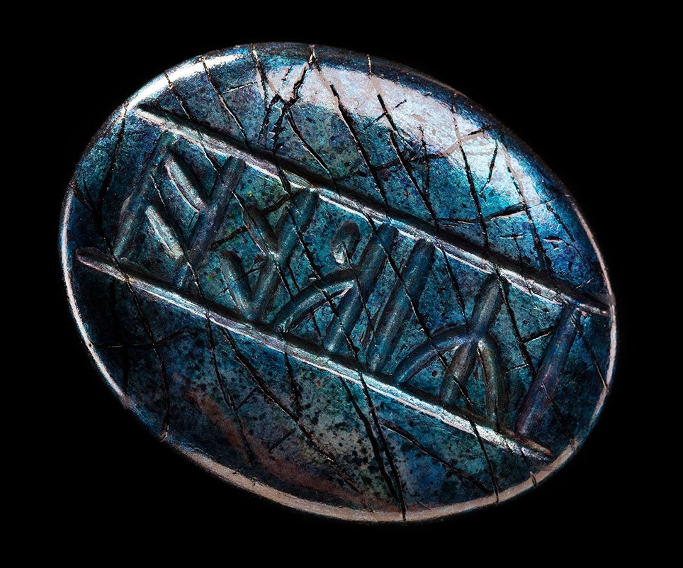 Kili's ovale runensteen, schuin afgebeeld op een zwarte achtergrond. Het glanzende, steenachtige oppervlak is blauwgroen met zilverkleurige randen, donkere kraslijnen en gegraveerde runen tussen twee diagonale groeven.